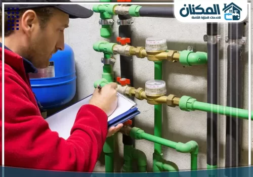سباك كشف تسربات بجدة 0569274510 فني بقميص أحمر يسجل قراءة عدادات المياه المتصلة بنظام أنابيب أخضر، لضمان الدقة في الاستهلاك.