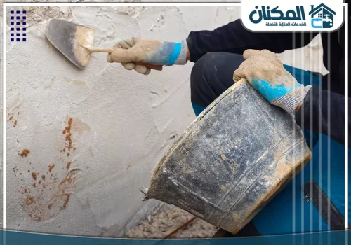 إصلاح تشققات الجدران مكة 0569274510 يد عامل بـقفازات تضع مونة اللياسة الطازجة على الجدار الخشن باستخدام مسطرين لتسوية السطح.